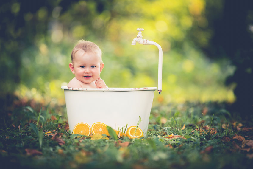 Baby sitzt lächelnd in einer alten kleinen Badewanne im Freien, mit Orangen als Deko 