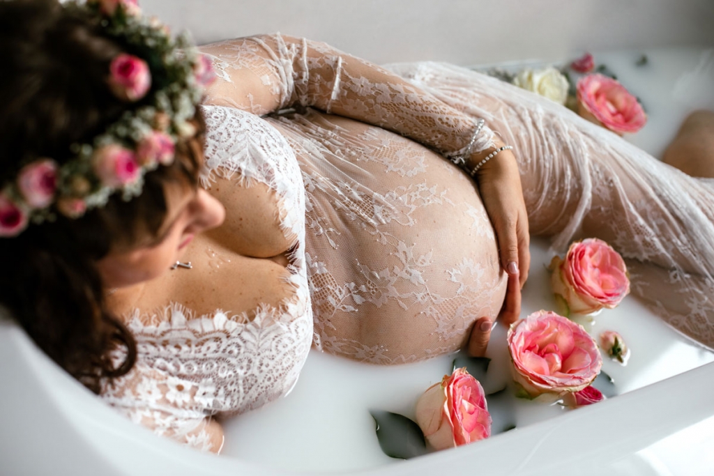 Junge Frau liegt seitlich in einem Milchbad, hält den Babybauch und ist von Blüten umgeben